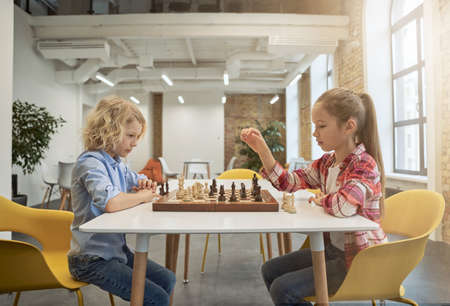 Two smart little children, boy and girl developing chess strategy, playing board game, sitting together at the table in schoolの写真素材