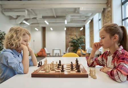 Chess matters. Clever children, boy and girl developing chess strategy, playing board game, sitting together at the table in schoolの写真素材