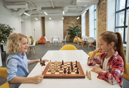 Two beautiful caucasian children, boy and girl smiling while playing chess, sitting together at the table in schoolの写真素材