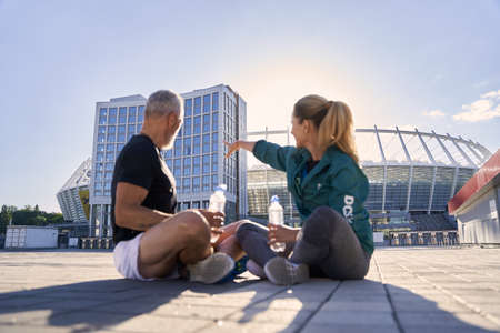 Active mature couple in sportswear discussing something while relaxing after workout, sitting outdoors on a sunny summer dayの写真素材
