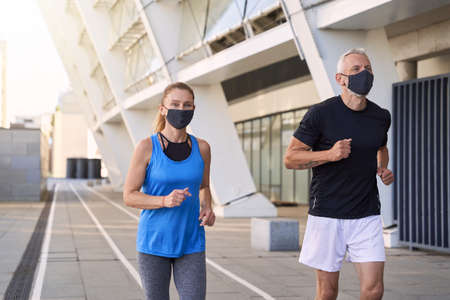 Sportive mature couple wearing protective face masks jogging together in urban environmentの写真素材