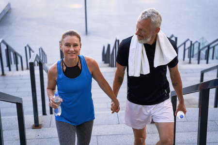 Attractive middle aged couple of sportive man and woman looking happy, holding hands while walking up the stairs after training together in the cityの写真素材