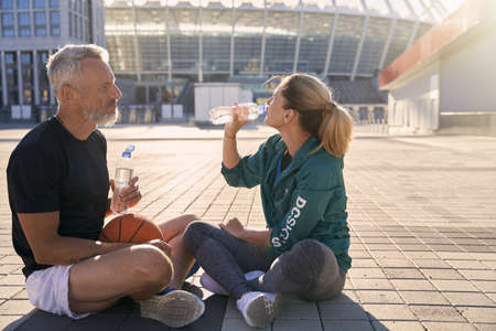 Two sportive friends, man and woman in sportswear drinking water while resting after workout, sitting outdoors on a sunny summer dayの写真素材