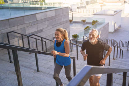 Mature couple in sportswear running up a flight of stairs in city while exercising together outdoors. Active sport, fitness, healthy lifestyle of middle aged peopleの写真素材