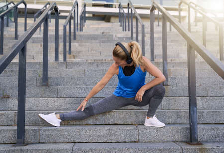 Fit middle aged woman in headphones stretching legs while working out outdoors in the cityの写真素材