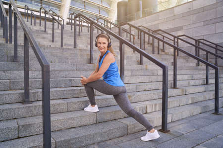 Full length shot of smiling middle aged woman in headphones warming up, exercising outdoors in the cityの写真素材