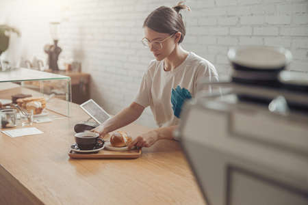 Charming young woman preparing coffee and bun for customer in cafeの写真素材