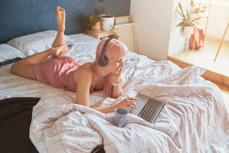 Beautiful woman with headphones surfs internet with laptop lying on bed in morningの写真素材