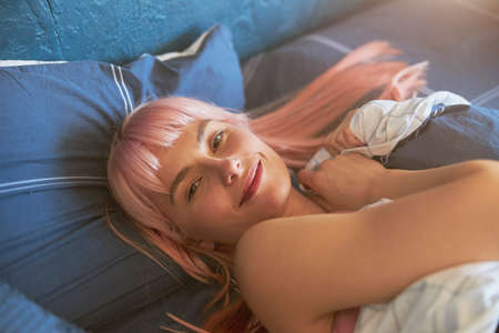 Portrait of attractive young woman with pink hair awakening in large comfortable bedの写真素材