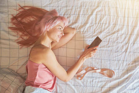 Smiling woman looks at mobile phone lying on comfortable bed upper viewの写真素材