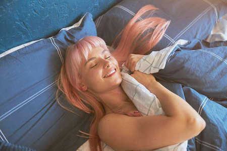 Smiling lady with stylish pink hair rests in large bed in lazy morningの写真素材