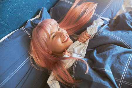 Positive female model with bright pink hair lies in large bed in early morningの写真素材