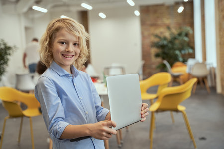 Portrait of excited little boy smiling at camera while holding and using laptop, standing in a classroom during STEM lessonの写真素材