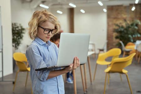 The future of IT. Portrait of little genius boy in glasses holding and using laptop while posing in a classroomの写真素材