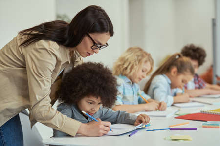 Attentive young female teacher helping little schoolboy. Kids sitting at the table, studying in elementary school classroomの写真素材