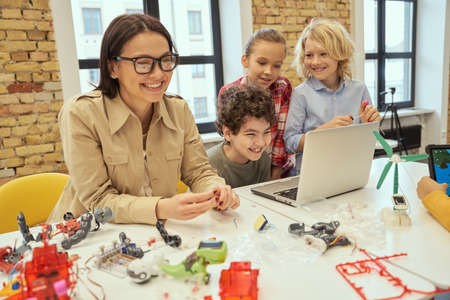 Joyful kids laughing together with their young female teacher while learning how to assemble robot using laptop, sitting at the table during STEM classの写真素材