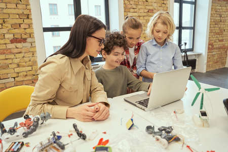 Be curious. Smiling kids watching scientific robotics video, sitting at the table in a classroom together with young female teacher during STEM classの写真素材
