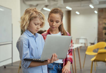 Portrait of little boy and girl holding and looking at laptop screen while standing in a classroom during STEM lessonの写真素材