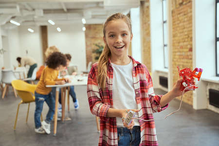 Portrait of excited little girl smiling at camera and showing her diy robot while standing in a classroom during STEM lessonの写真素材