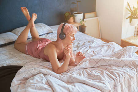 Stylish woman wearing pink silk pajamas enjoys with headphones lying on comfortable bedの写真素材