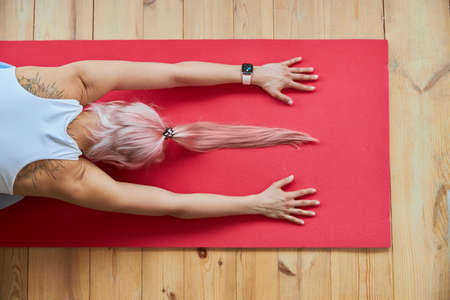 Pink haired lady with smartwatch in white top sits in child pose on floorの写真素材
