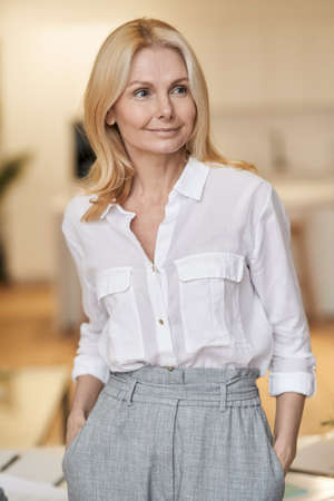 Elegant mature business woman with blonde hair looking aside while standing indoorsの写真素材