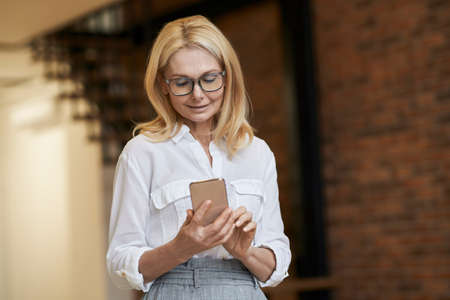 Technology. Beautiful mature business woman with blonde hair and glasses using her smartphone, standing indoorsの写真素材