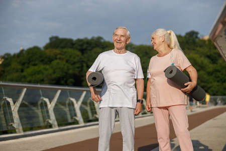 Cheerful mature couple with rolled mats ready for workout on footbridgeの写真素材