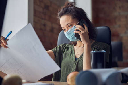 Portrait of female interior designer or architect wearing protective mask talking on the phone, dicussing a blueprint while working on new poject, sitting at the table in her officeの写真素材