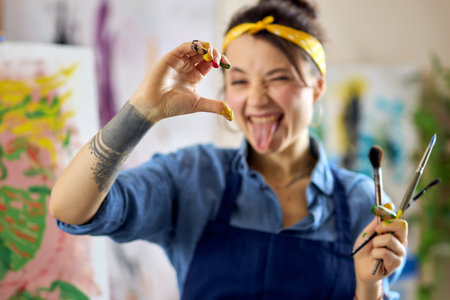 Playful female artist showing half heart love sign gesture with her hand in paint at camera while...