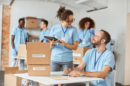 Man and woman happy to work together on donation project indoors, calculating, sorting and packing items in cardboard boxesの写真素材