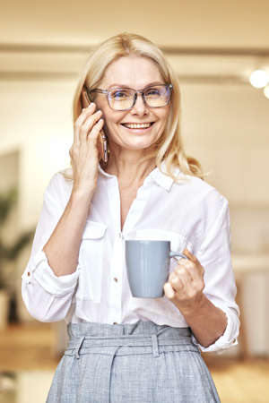 Portrait of attractive mature blonde businesswoman smiling at camera while making call and drinking coffee, standing in her officeの写真素材
