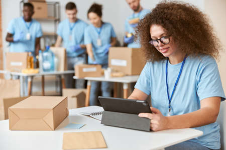 Attractive young woman, volunteer in blue uniform using tablet pc while sitting indoors, working on donation project. Team sorting, packing items in the backgroundの写真素材
