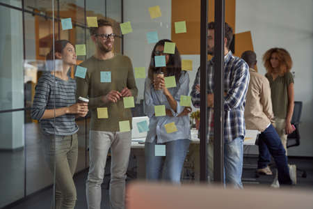 Succesful team. Group of happy business people, coworkers looking at sticky notes on a glass board, discussing new project and drinking coffee, working together in the officeの写真素材