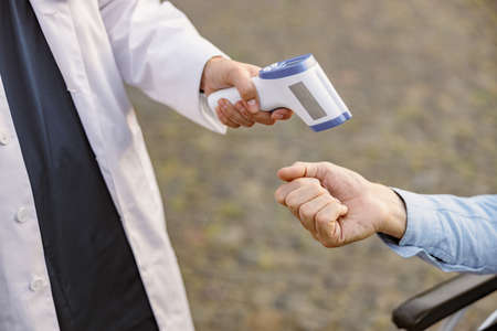 Close up of medical infrared thermometer in a hand of doctor measuring the temperature of male handicapped patient in wheelchair during outbreakの写真素材