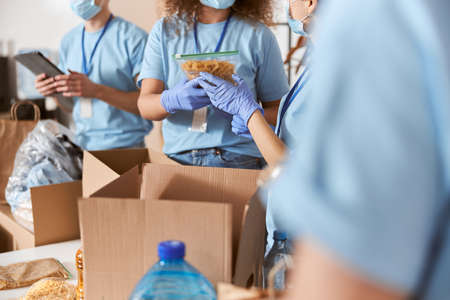 Close up shot of busy volunteers wearing blue uniform, protective masks and gloves sorting donated pack of pasta and other food items while working in charitable foundationの写真素材