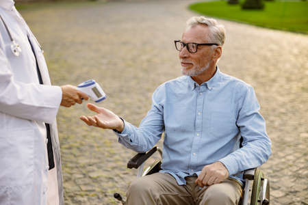 Senior handicapped man reaching out hand for doctor to check fever by digital thermometer. Scanning and protectionの写真素材