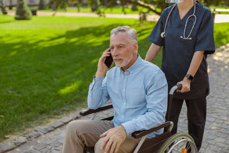Senior man, recovering patient in wheelchair talking on the phone while walking with nurse in the park on a summer dayの写真素材