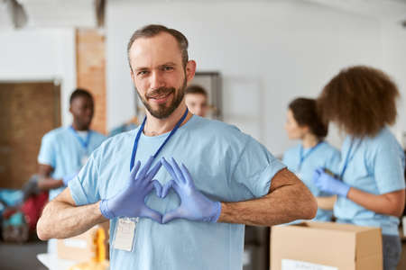 Portrait of caucasian male volunteer in blue uniform and protective gloves showing heart love sign, looking at camera. Team sorting, packing items in cardboard boxesの写真素材