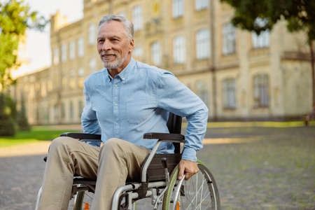 Smiling aged recovering man in wheelchair looking away while spending time alone in the park near hospitalの写真素材