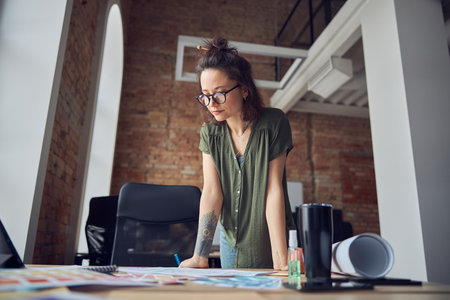 Creative woman, interior designer or architect in casual wear with messy hairdo looking at blueprint while working on new project, standing in her officeの写真素材