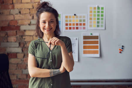 Smiling woman, interior designer in casual wear with messy hairdo looking at camera, holding glasses, standing in front of board with color samples while working on new creative projectの写真素材