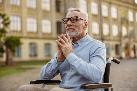 Portrait of mature handicapped man in wheelchair looking thoughtful while spending time alone outdoors near rehabilitation clinicの写真素材