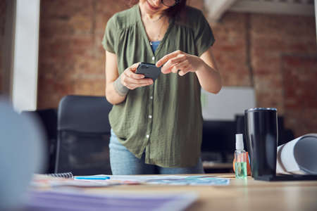 Creative woman, interior designer or architect texting or taking picture of a blueprint using smartphone, working on new project, standing in her office. Focus on phoneの写真素材