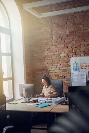 Concentrated woman in glasses, professional architect or designer working on blueprint, sitting at the table in her officeの写真素材