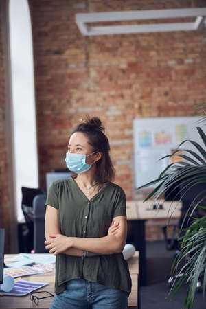Creative business woman in casual wear wearing protective mask looking aside and leaning on the table while standing in modern green office on a daytimeの写真素材