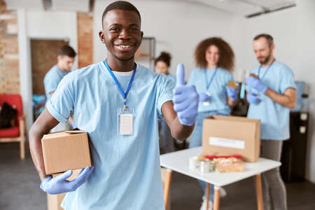Portrait of african american young male volunteer in blue uniform, protective gloves smiling at camera, showing thumbs up and holding cardboard box for Donationの写真素材