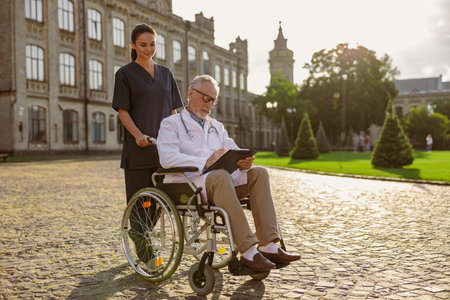 Full length shot of senior handicapped male doctor in wheelchair wearing lab coat and glasses with young nurse assisting him outdoorsの写真素材