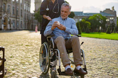 Focused senior man recovering patient holding cream jar, listening to nurse advice, walking together outdoors near rehabilitation clinicの写真素材