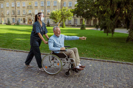Full length shot of caring nurse wearing face shield and mask spending time with senior male patient in wheelchair on a walk in the park near hospitalの写真素材